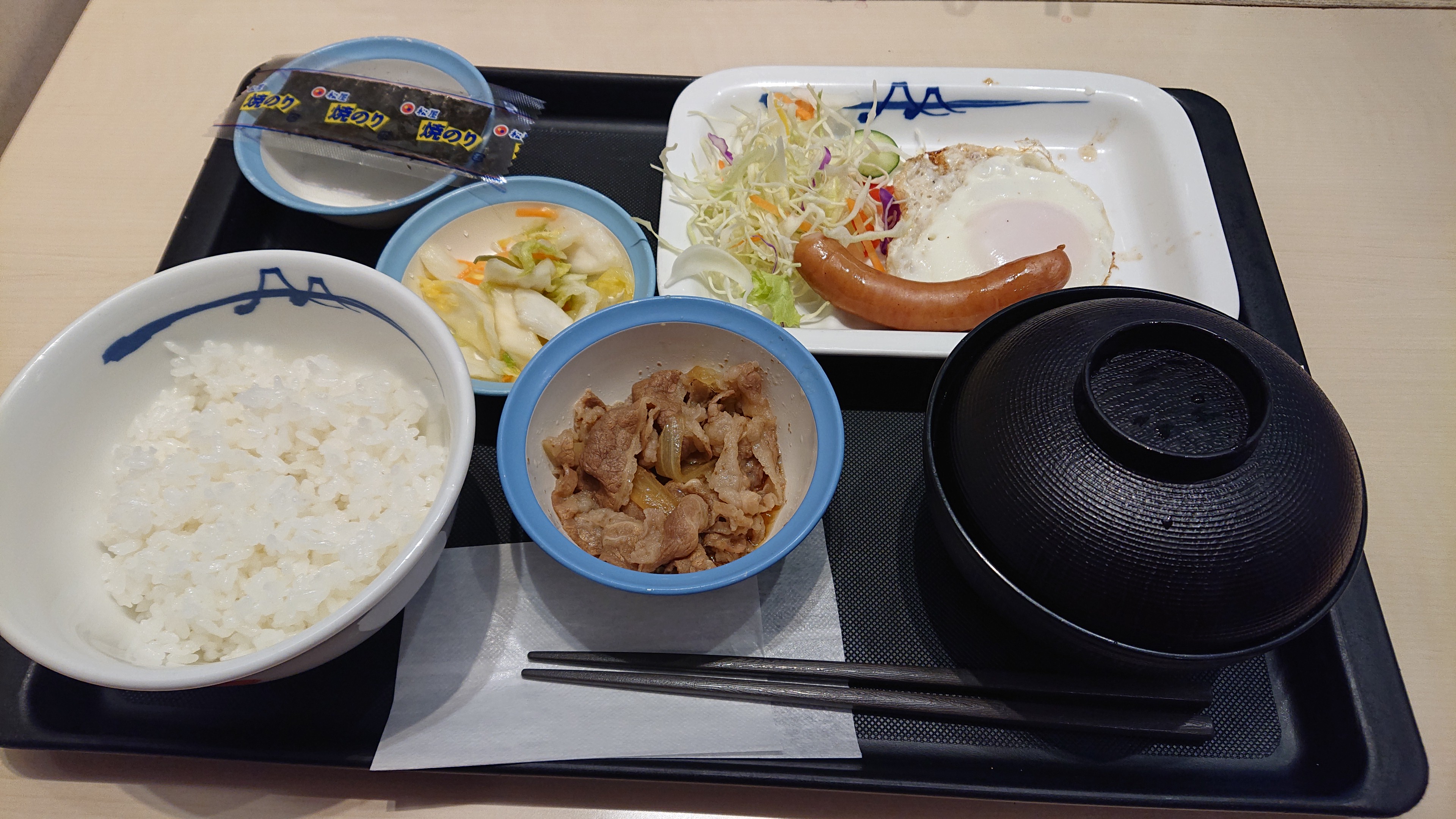 松屋で提供された、ごはん・目玉焼き・ソーセージ・牛肉の小鉢などが並ぶ朝食の参考写真